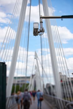 Detail of surveillance camera with unrecognisable people walking by in blurred background, and blue sky above.