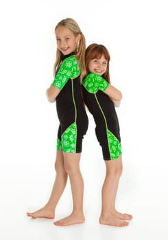 Vertical photo of two happy, Caucasian little girls standing back to back wearing wetsuits on white background
