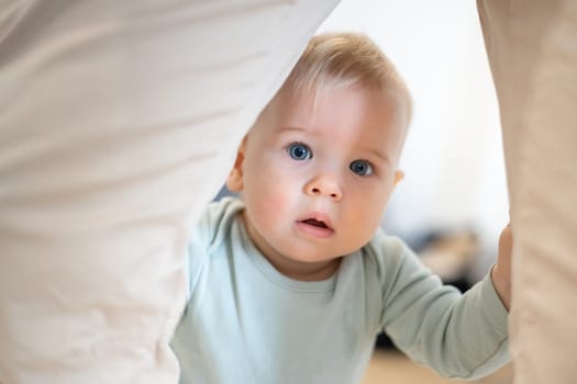 Portrait of adorable curious infant baby boy child taking first steps holding to father's pants at home. Cute baby boy learning to walk