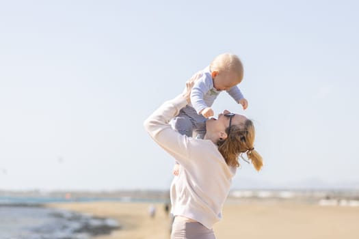 Mother enjoying summer vacations holding, playing and lifting his infant baby boy son high in the air on sandy beach on Lanzarote island, Spain. Family travel and vacations concept