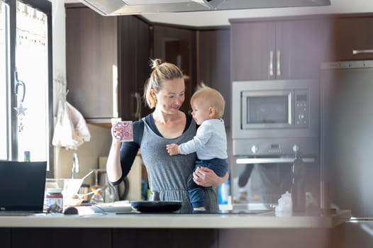 Happy mother holding her little infant baby boy while drinking morning coffee and making pancakes for breakfast in domestic kitchen. Family lifestyle, domestic life concept