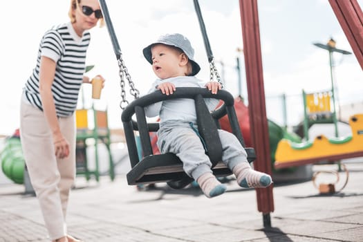 Mother pushing her infant baby boy child on a swing on playground outdoors.