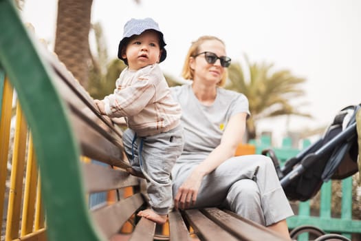 Young mother with her cute infant baby boy child on bench on urban children's playground on warm summer day