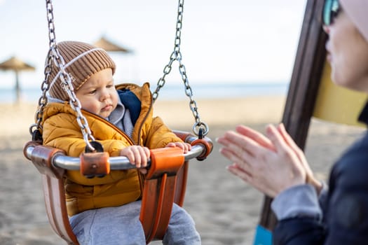 Mother pushing her infant baby boy child on a swing on sandy beach playground outdoors on nice sunny cold winter day in Malaga, Spain