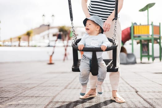 Mother pushing her infant baby boy child on a swing on playground outdoors