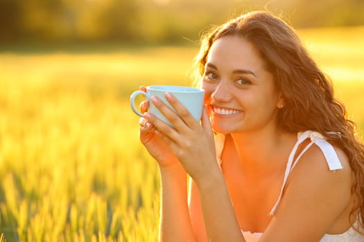 Happy woman in a field looking at you drinking coffee at sunset