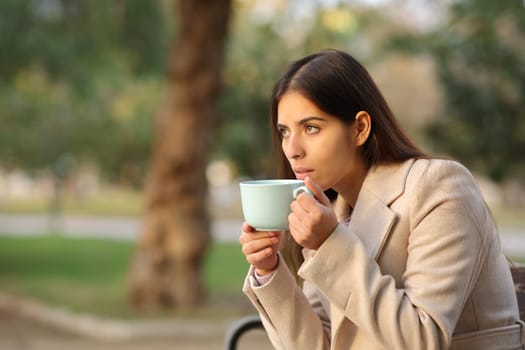 Woman drinking coffee looks away at sunset in a park in winter