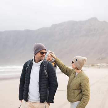 Young father carrying his infant baby boy son in backpack on windy sandy Famara beach, Lanzarote island, Spain. Family travel and winter vacation concept