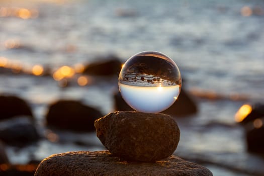 Glass ball lies on the top stone of stacked stones, at sunset on the beach, the sea and the setting sun are reflected in the ball