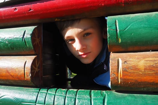 A 10-year-old Caucasian boy with blond hair and gray eyes looks out of the window of a wooden house on a children's playground. Peekaboo. The child hid. Pretty face.