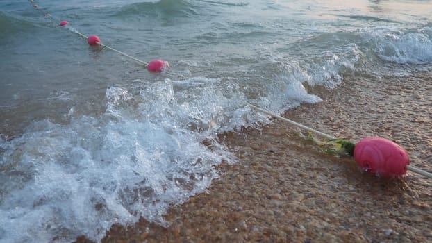 Buoys on a rope near sea water. The buoys are pink restraints to alert people to the depth of the water. Rescue of the drowning. Delimiting a place on a sandy beach between hotels. Wave with bubbles