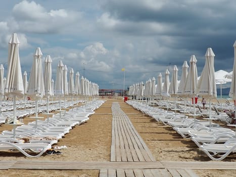 Wet sunbeds and umbrellas on the sea beach during heavy rain storm. Rolled up parasol on the beach against dark cloudy sky. Invasion of a typhoon, cyclone or storm on the sea or ocean. Beach season