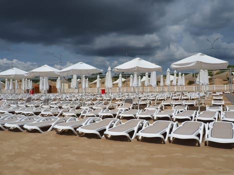 Wet sunbeds and umbrellas on the sea beach during heavy rain storm. Rolled up parasol on the beach against dark cloudy sky. Invasion of a typhoon, cyclone or storm on the sea or ocean. Beach season
