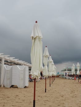 Wet sunbeds and umbrellas on the sea beach during heavy rain storm. Rolled up parasol on the beach against dark cloudy sky. Invasion of a typhoon, cyclone or storm on the sea or ocean. Beach season.