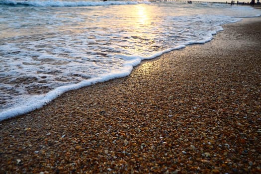 Foamy waves roll over the sandy shore. Sunset over the sea. Beautiful sunset. Tourist place, resort. The sun's rays are reflected from the water. Sandy evening beach with seashells at golden hour.