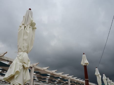 Wet sunbeds and umbrellas on the sea beach during heavy rain storm. Rolled up parasol on the beach against dark cloudy sky. Invasion of a typhoon, cyclone or storm on the sea or ocean. Beach season.