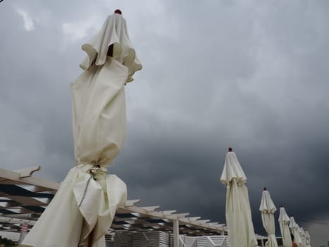 Wet sunbeds and umbrellas on the sea beach during heavy rain storm. Rolled up parasol on the beach against dark cloudy sky. Invasion of a typhoon, cyclone or storm on the sea or ocean. Beach season.