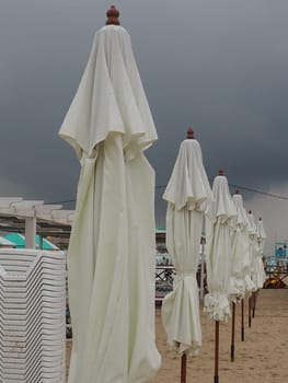Wet sunbeds and umbrellas on the sea beach during heavy rain storm. Rolled up parasol on the beach against dark cloudy sky. Invasion of a typhoon, cyclone or storm on the sea or ocean. Beach season.