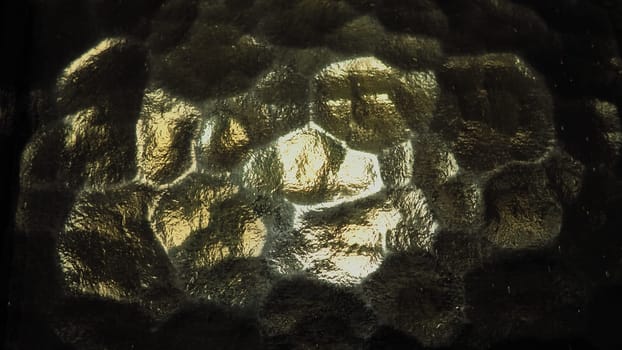 Corrugated glass, rear light source. Play of glare and reflections on a transparent uneven surface. Dark and light abstract asymmetric multi-illuminated planes. Abstract bumpy golden background