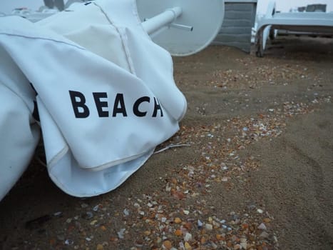 A beach umbrella lay folded on a sandy beach after bad weather. Storm at sea. End of the beach season due to typhoon and cyclone. Loungers, umbrellas and hotel furniture. Beach word written on white.