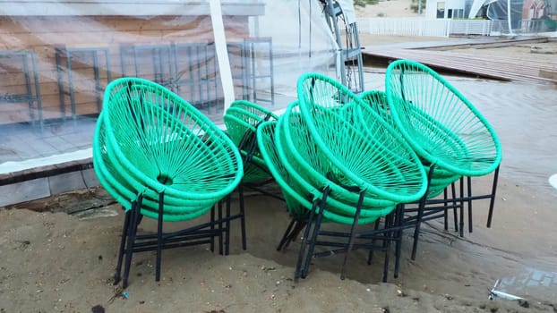 wicker green plastic chairs with black legs in a cafe on a sandy beach after a storm. The furniture is stacked on top of each other as the beach cafe is closed. End of the beach season at the hotel.