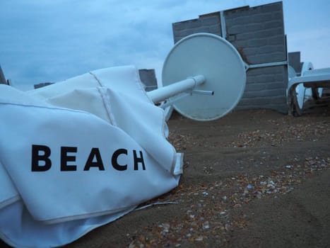 A beach umbrella lay folded on a sandy beach after bad weather. Storm at sea. End of the beach season due to typhoon and cyclone. Loungers, umbrellas and hotel furniture. Beach word written on white.
