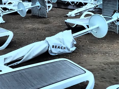 A beach umbrella lay folded on a sandy beach after bad weather. Storm at sea. End of the beach season due to typhoon and cyclone. Loungers, umbrellas and hotel furniture. Beach word written on white.
