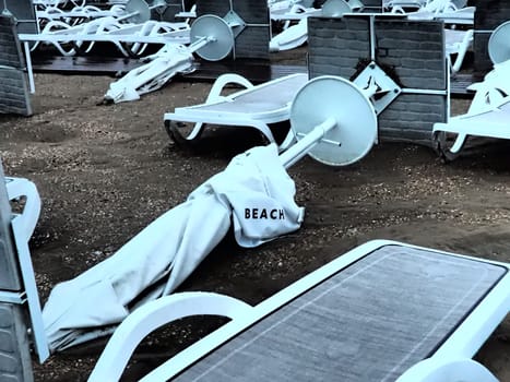 A beach umbrella lay folded on a sandy beach after bad weather. Storm at sea. End of the beach season due to typhoon and cyclone. Loungers, umbrellas and hotel furniture. Beach word written on white