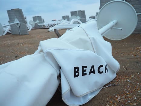 A beach umbrella lay folded on a sandy beach after bad weather. Storm at sea. End of the beach season due to typhoon and cyclone. Loungers, umbrellas and hotel furniture. Beach word written on white.