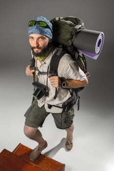 Portrait of a male fully equipped tourist  climbing up the stairs with backpack and the camera on gray background
