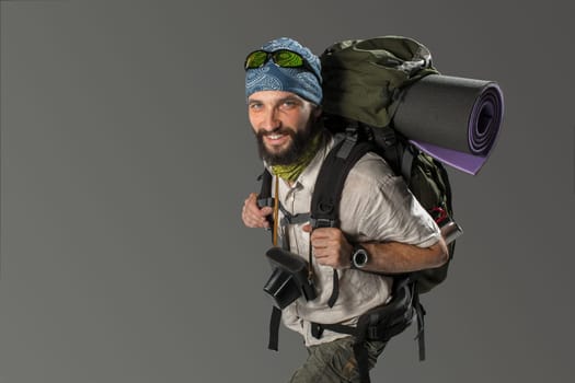 Portrait of a male smiling fully equipped tourist with backpack and the camera on gray background