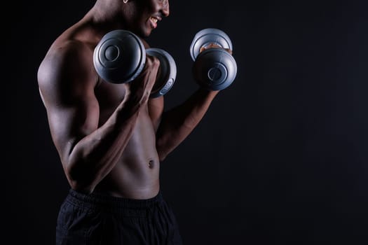 Confident young man shirtless portrait training with dumb-bell against black background.