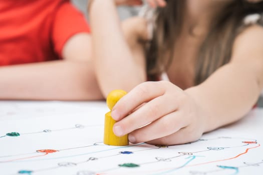 Close up child hand with yellow game paw figure on creative art map. Girl and other kids playing board game with pieces. Family fun. Educational strategy for whole family. Strategic game development