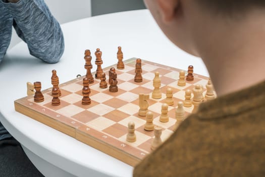 Wooden chess battle on chess board on white table between unrecognizable rearview of boys. Strategic game, training brain skills, preparing for competition. Leadership and genius kids in logical games
