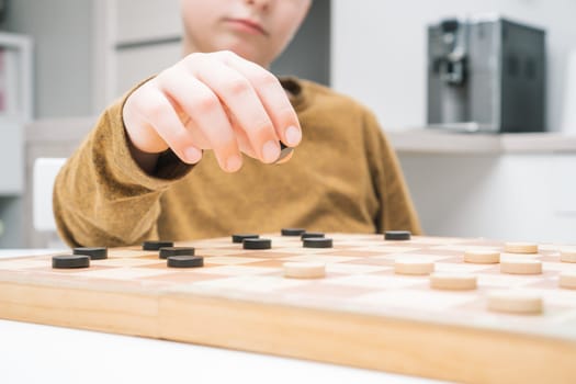 Close up unrecognizable cropped little boy hand with checker piece making step on wooden board in kitchen. Strategy and logic skills with educational games. Children playing smart board games