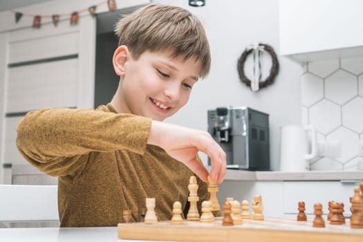 Smiling little boy joying playing chess, looking at king piece on wooden board on table in kitchen. Intelligence and smart activity. Strategic game, training brains. Young grand chess master in work