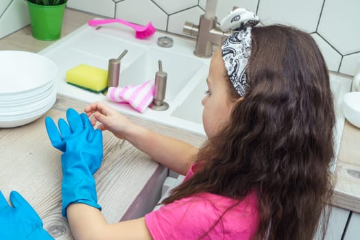 Little girl taking off blue household gloves after washing up dish. Portrait of child washing tableware in kitchen sink side view. Home cleaning concept. Personal protective equipment.