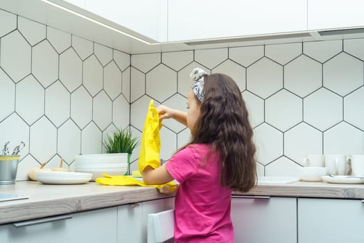Little girl taking off yellow household gloves after washing up dish. Portrait of child washing tableware in kitchen sink back view. Home cleaning concept. Personal protective equipment.