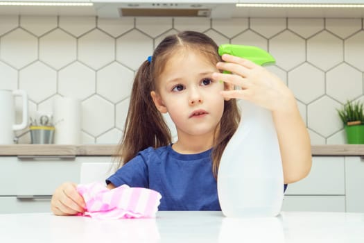 Pretty little girl push lever of detergent sprayer at table. Portrait of child tidying up kitchen with household rag, home cleaning concept. Kid help clean up house. Room cleaning supplies, cleanser.