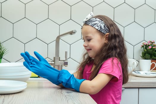 Happy little girl putting on blue household gloves for washing up dish. Portrait of child washing tableware in kitchen sink side view. Home cleaning concept. Personal protective equipment.