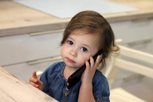Cute little dark-haired girl 1,5-2,5 in a jeans dress is talking to someone on a smartphone in the kitchen. Baby miss communication with relatives. Self-isolation and quarantine concept.