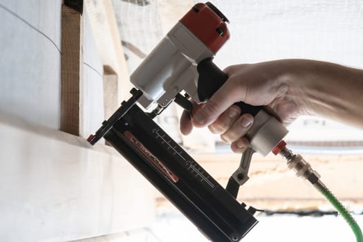 Cropped man hand hold pneumatic stapler nail gun for wooden timber plank. Clipping and scoring metal nails and staples, making layout on wooden framework. Builder occupation, civil engineer, Side view