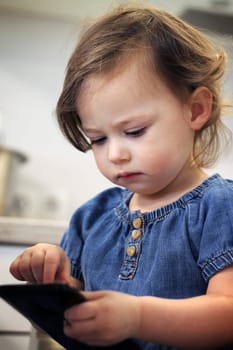 Cute little dark-haired girl 1.5-2.5 in a jeans dress types something or dials a number on a smartphone in the kitchen. Baby miss communication with relatives. Self-isolation and quarantine concept.