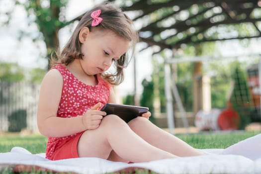 Little 3-4 girl in red clothes sits on blanket on green grass and looks into mobile phone. Children using gadgets