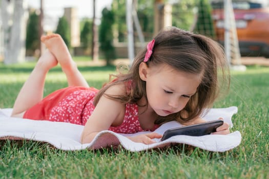 Little 3-4 girl in red clothes lies on blanket on green grass and looks into mobile phone. Children using gadgets