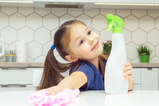 Nice little girl with detergent sprayer and household rag wipe table. Portrait of happy child tidying up kitchen, home cleaning concept. Kid help clean up house. Room cleaning supplies, cleanser.