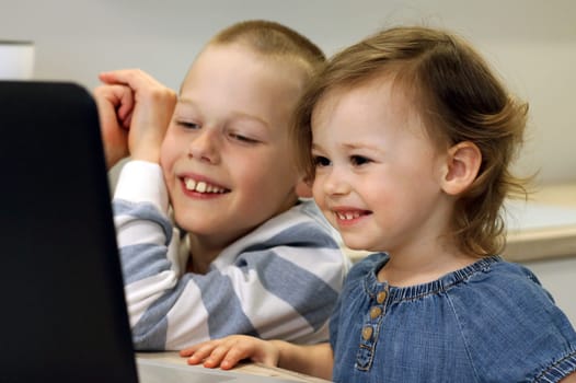 Two laughing children are sitting in the kitchen and looking at the computer. Children look at a laptop and communicate with their relatives online using the Internet. Self-isolation concept.