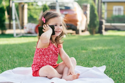 Little 3-4 girl in red clothes sits on blanket on green grass and speaks on cell phone. Children using gadgets