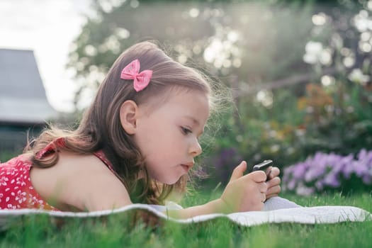 Portrait of little girl 3-4 in red clothes lying on blanket on green grass and looking into mobile phone. Children using gadgets