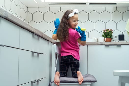 Happy little girl in protective gloves wash up dishes with sponge in kitchen sink sitting on chair. Portrait of child tidying up kitchen back view. Home cleaning concept. Stylish interior, furniture.